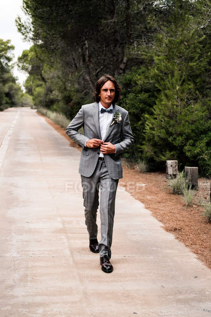 Jovem noivo em elegante terno de casamento cinza indo em estrada vazia com árvores verdes em lados — Fotografia de Stock