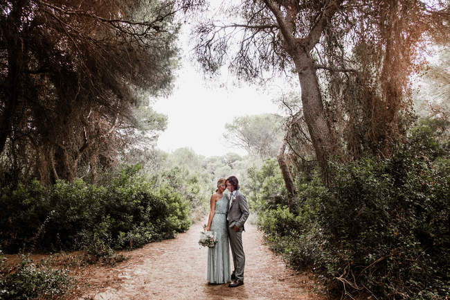 Junges frischvermähltes Paar in Hochzeitskleidung steht und umarmt auf dem Weg zwischen wunderschönem grünen Wald mit großen Bäumen — Stockfoto