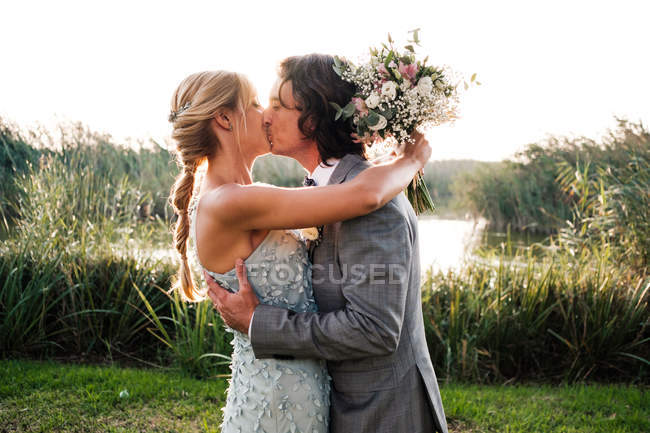 Stylisches frisch verheiratetes Paar in Hochzeitsanzügen umarmt und küsst sich mit grünen Pflanzen und nur an einem sonnigen Tag — Stockfoto