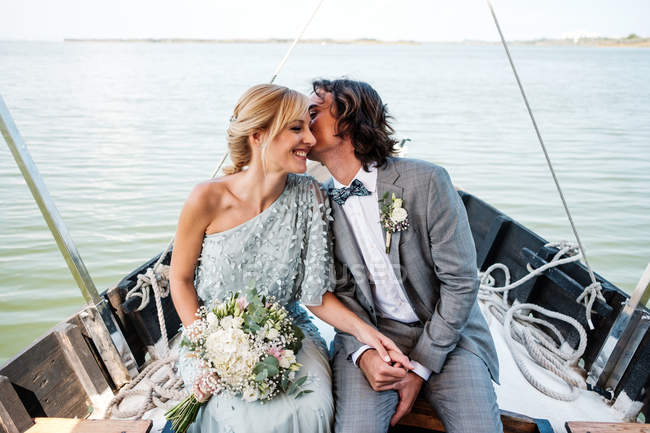 Satisfied married lovers in wedding clothing kissing while relaxing on boat with sea on background — Stock Photo
