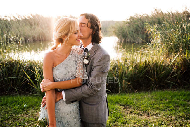 Elegante coppia di sposi in abiti da sposa abbracciare e baciare con piante verdi e mera giornata di sole — Foto stock