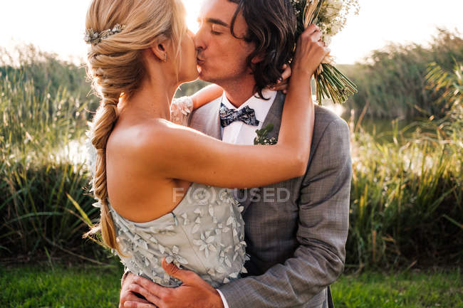 Stylish newly married couple in wedded suits embracing and kissing with green plants and mere on sunny day — Stock Photo