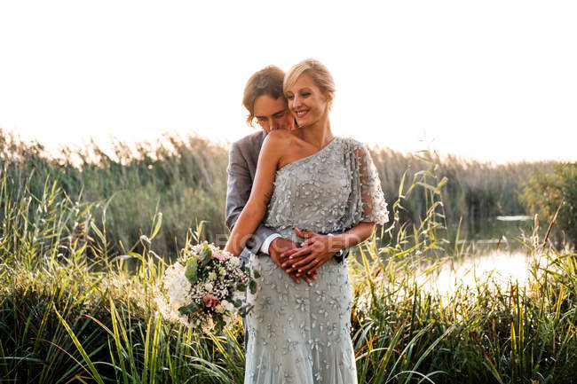 Стильна нещодавно одружена пара у весільних костюмах, яка обіймає і цілує зеленими рослинами і просто в сонячний день — стокове фото