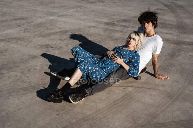 Mulher loira sensual atraente olhando para a câmera, deitada no homem sentado no skate na praça — Fotografia de Stock