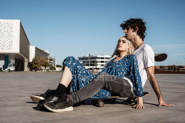 Attraente donna bionda sensuale con gli occhi chiusi sdraiati sull'uomo seduto sullo skateboard e sognando contro il cielo blu — Foto stock