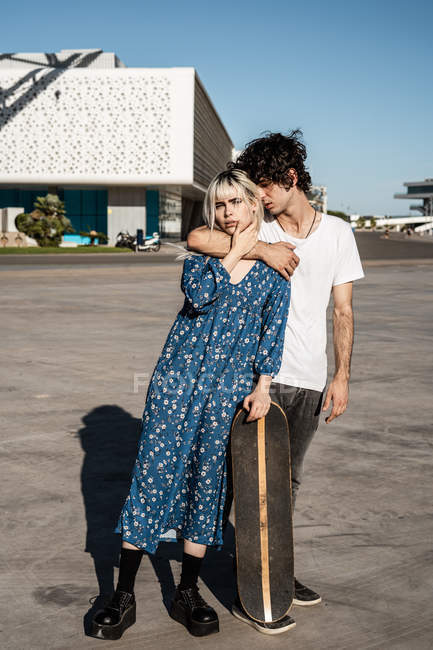 Молодая модная любящая пара, стоящая и опирающаяся на скейтборд на площади перед голубым небом и размытые современные здания — стоковое фото