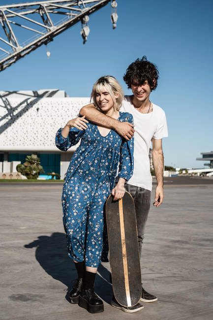 Молода модна любляча пара стоїть і спирається на скейтборд на площі проти блакитного неба і розмитих сучасних будівель — стокове фото