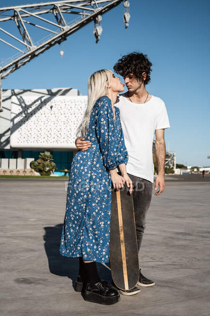 Jeune couple aimant tendance debout et appuyé sur le skateboard sur la place contre le ciel bleu et les bâtiments modernes flous — Photo de stock