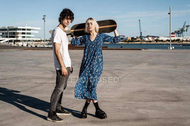 Молодая модная любящая пара стоит, пока женщина держит скейтборд на площади против голубого неба и размытых современных зданий — стоковое фото