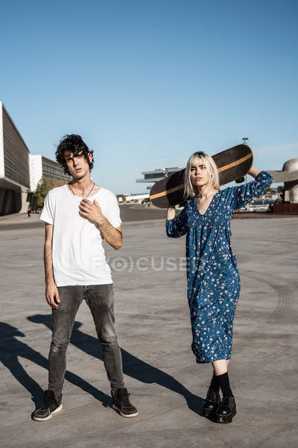 Junges trendiges Liebespaar steht, während Frau ein Skateboard auf dem Platz vor blauem Himmel und verschwommenen modernen Gebäuden hält — Stockfoto