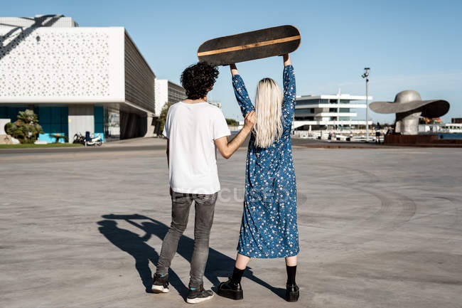 Rückansicht eines jungen trendigen Pärchens, das steht, während Frau ein Skateboard gegen blauen Himmel auf den Platz hebt — Stockfoto