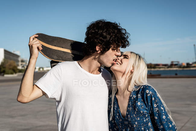 Attractive young stylish couple embracing and kissing each other on the street while man in white shirt holding skateboard — Stock Photo