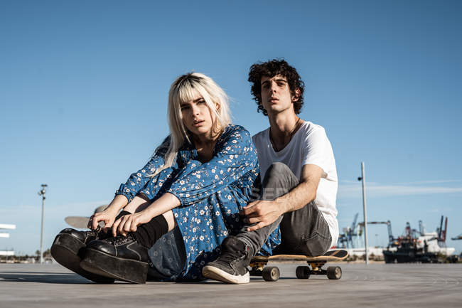 Attraente sensuale donna bionda guardando la fotocamera seduta con il fidanzato sullo skateboard e sognando contro il cielo blu — Foto stock