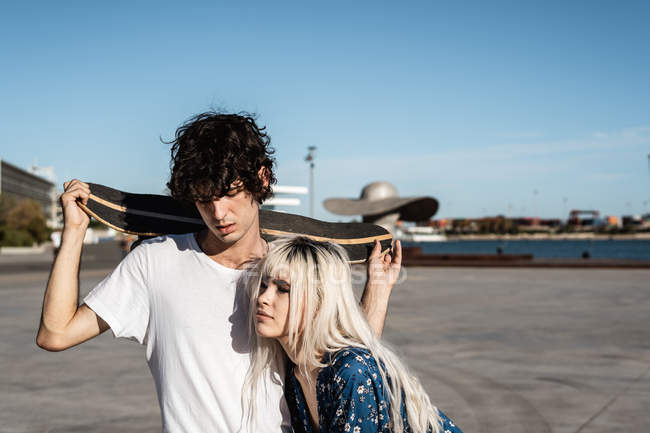 Attraente sensuale donna bionda con gli occhi chiusi abbracciando giovane uomo dai capelli scuri in camicia bianca con lo skateboard in mano — Foto stock