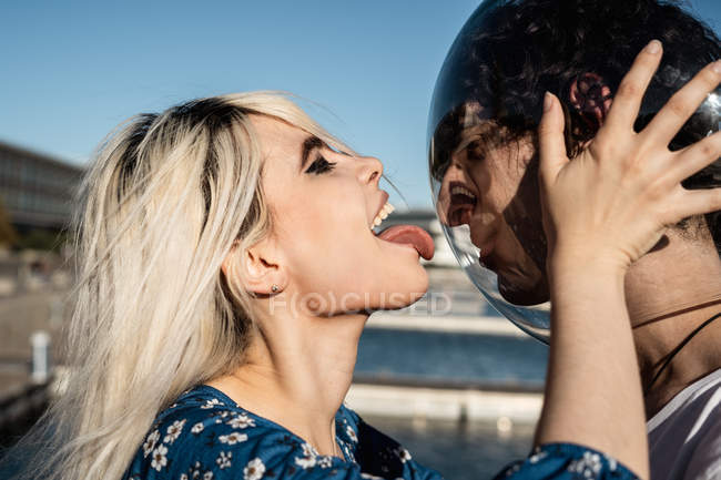 Side view of attractive blond female with closed eyes licking transparent round aquarium on head of adult man — Stock Photo