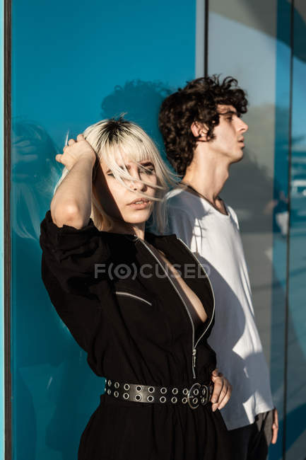 Женщина в черной одежде стоит рядом с высоким мужчиной с закрытыми глазами, стоя напротив голубой стены с отражением улицы — стоковое фото