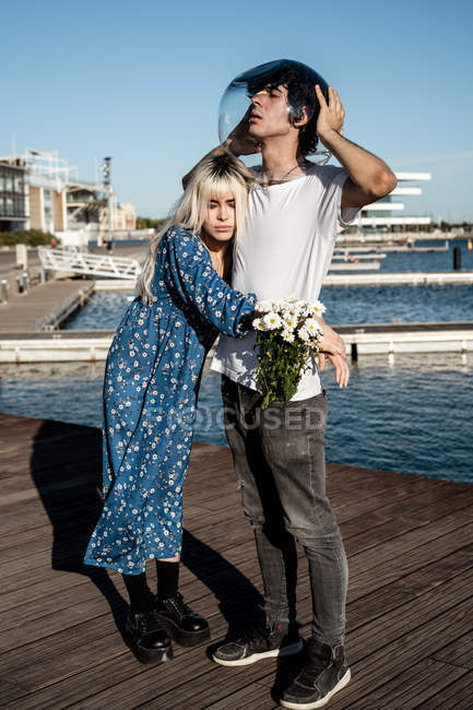 Attractive unhappy blond female with bouquet of daisies hugging man with transparent round aquarium on head — Stock Photo
