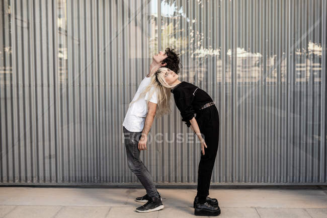 Vista lateral de la pareja de moda apoyándose mutuamente mientras están de pie espalda con espalda y soñando contra la pared rayada - foto de stock