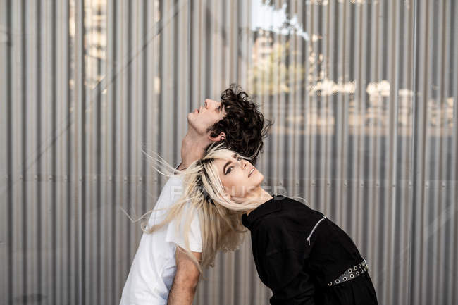 Vista lateral de la pareja de moda apoyándose mutuamente mientras se paran espalda con espalda contra la pared rayada del edificio moderno - foto de stock
