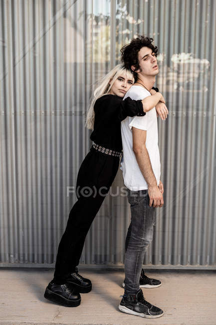 Seitenansicht des trendigen Paares, das sich gegenseitig stützt, während sich die Frau auf dem Rücken ihres Freundes gegen die gestreifte Wand des modernen Gebäudes lehnt — Stockfoto