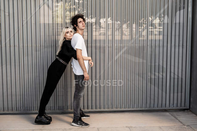 Vista lateral do casal na moda apoiando uns aos outros, enquanto a mulher se inclina no namorado de volta contra a parede listrada do edifício moderno — Fotografia de Stock