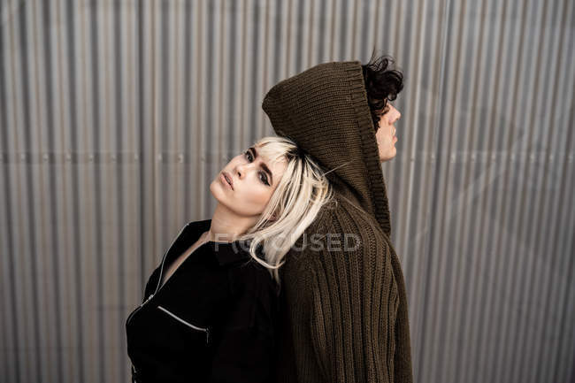Vista lateral do casal na moda apoiando uns aos outros enquanto estão de costas para trás e sonhando contra a parede listrada — Fotografia de Stock