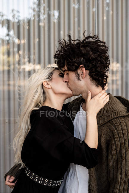 Seitenansicht einer Frau, die nach Zunge beißt und den Hals eines jungen lockigen dunkelhaarigen Mannes berührt, während er steht und küsst — Stockfoto