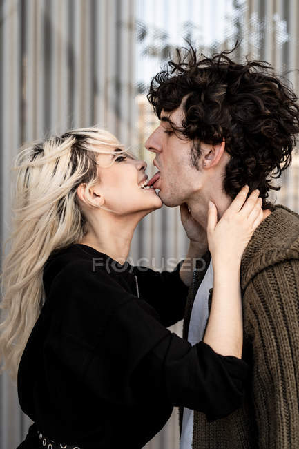 Donna bionda adulta che morde per la lingua e tocca il collo del giovane uomo dai capelli ricci scuri mentre sta in piedi e bacia — Foto stock