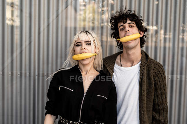 Extraordinario hombre y mujer de moda sosteniendo plátanos en la boca sobre el fondo de la pared rayada - foto de stock
