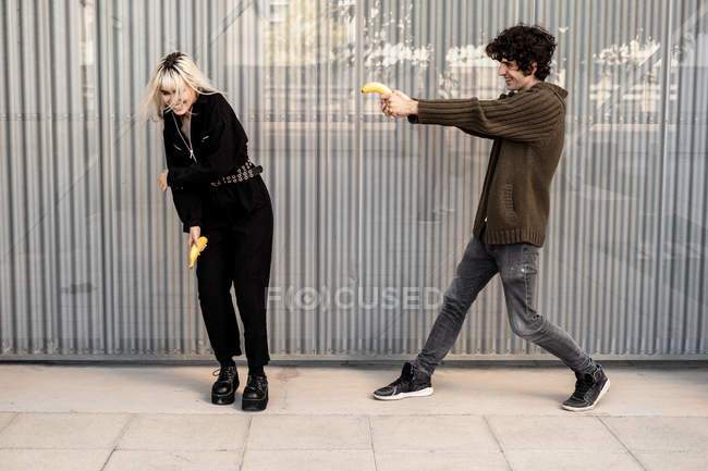 Cheerful man aiming with banana at woman protecting with hands and laughing while having fun together on street — Stock Photo