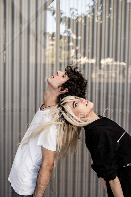 Seitenansicht des trendigen Paares, das sich gegenseitig stützt, während es Rücken an Rücken gegen die gestreifte Wand des modernen Gebäudes steht — Stockfoto