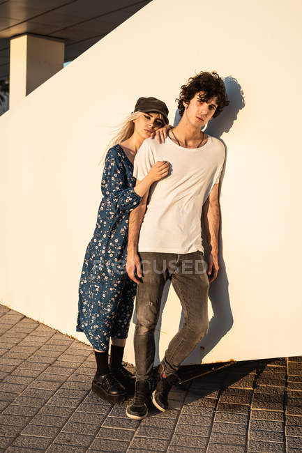 Junges stylisches Paar umarmt einander, während es sonnigen Tag genießt und betrachtet, an weiße Wand gelehnt — Stockfoto