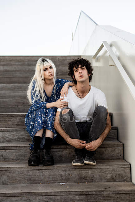 Модная взрослая женщина и мужчина в повседневной одежде сидят на деревянной лестнице, смотрят в камеру — стоковое фото