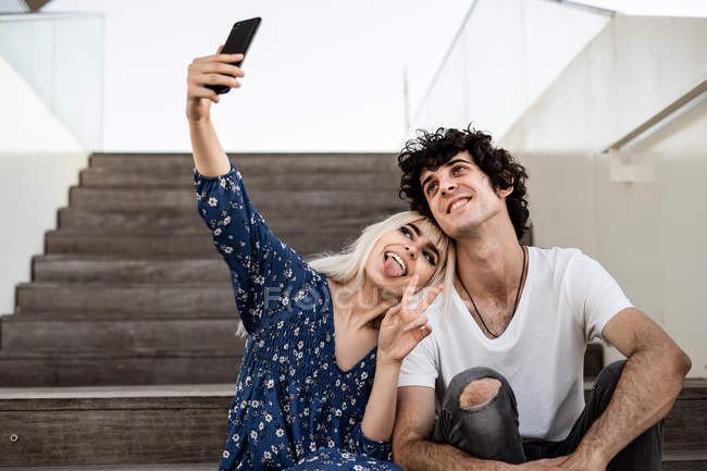 Smiling trendy adult woman and man in casual clothes taking selfie together with smartphone while sitting on wooden stairs — Stock Photo