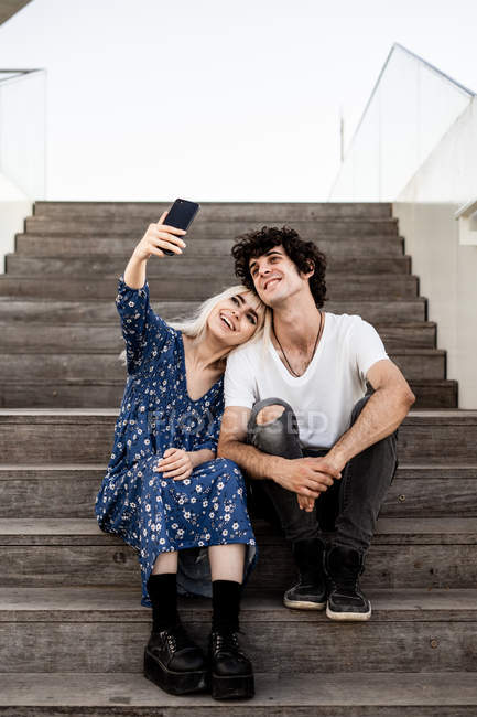 Усміхнена модна доросла жінка і чоловік в повсякденному одязі беруть селфі разом зі смартфоном, сидячи на дерев'яних сходах — стокове фото