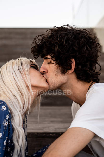 Vista lateral del hombre de pelo oscuro rizado adulto con ojos cerrados besando a la mujer rubia contra escaleras borrosas a la luz del día - foto de stock