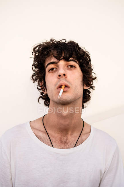 From below handsome confident dark haired man in casual clothes with cigarette in mouth looking with curiosity at camera — Stock Photo