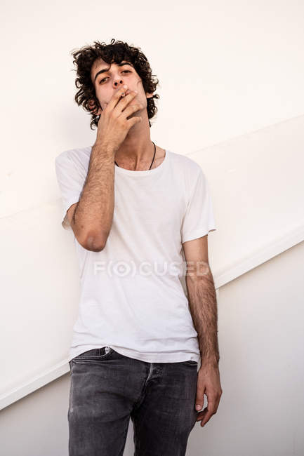 Знизу гарний, впевнений, темноволосий чоловік у звичайному одязі з сигаретою у роті, з цікавістю дивлячись на камеру. — стокове фото