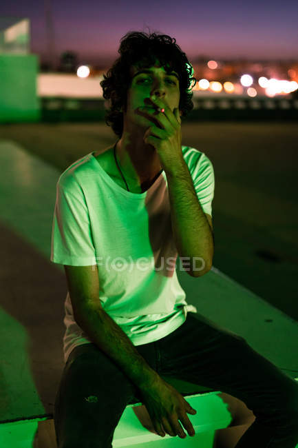 Красивый парень в повседневной одежде с сигаретой во рту, с интересом смотрит в камеру на размытом фоне улицы — стоковое фото