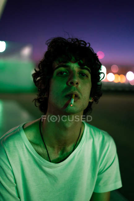 Beau gars en vêtements décontractés avec cigarette dans la bouche regardant avec intérêt la caméra sur fond flou de la rue — Photo de stock