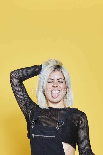 Привлекательная блондинка в черном комбинезоне, стоящая с закрытыми глазами и смешным лицом на желтом фоне — стоковое фото