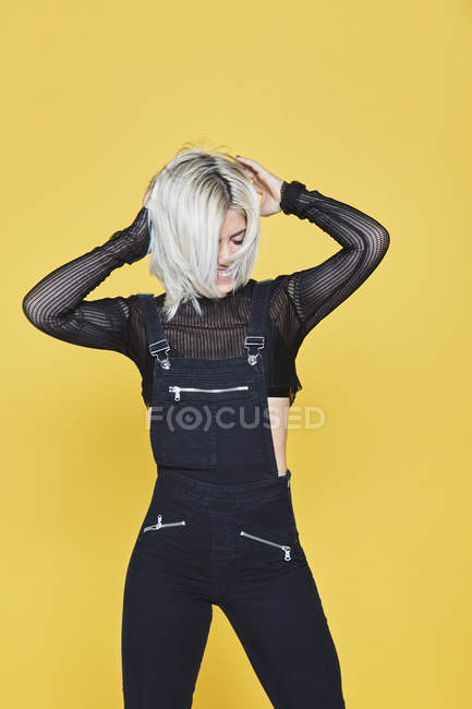Attraktive blonde Frau in schwarzen Overalls, die auf gelbem Hintergrund steht und nach unten schaut — Stockfoto