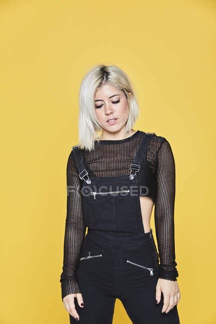 Attractive blonde woman in black overalls standing and looking down on yellow background — Stock Photo