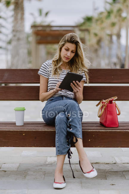 Женщина в полосатой рубашке и джинсах сидит на уличной скамейке у моря с одноразовой чашкой кофе и просматривает интернет на планшете — стоковое фото