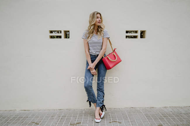 Mulher com bolsa de ombro vermelho vestindo camisa listrada e jeans de pé com as mãos dobradas e olhando para longe com sorriso tímido — Fotografia de Stock
