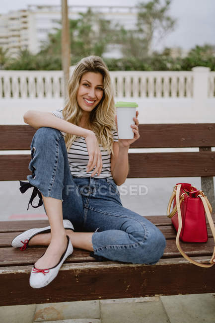 Femme gaie avec tasse de café à emporter assis sur le banc de la ville en bord de mer le jour de l'été — Photo de stock