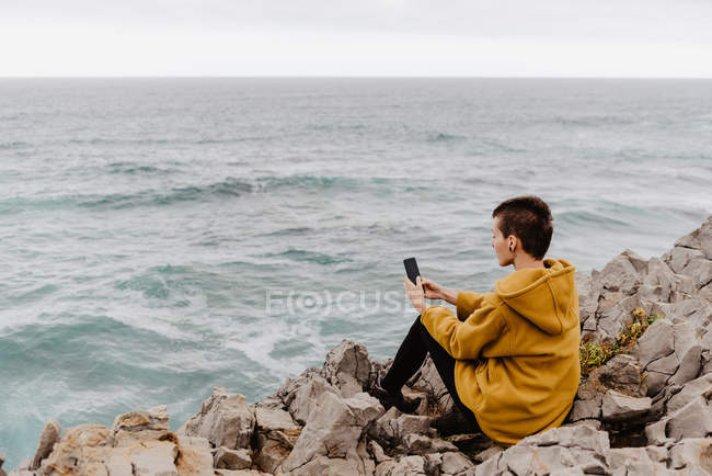 Back view of short haired woman in yellow sweatshirt sitting on rocky seashore and taking selfie on mobile phone — Stock Photo