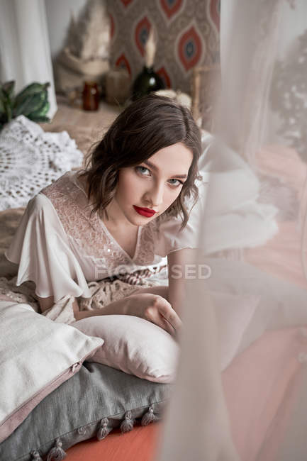 Mulher linda com lábios vermelhos em vestido branco olhando para a câmera enquanto sentado no chão ao lado do sofá — Fotografia de Stock