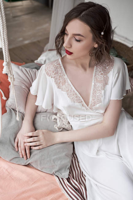 Великолепная женщина с красными губами в белом платье смотрит в сторону, сидя на полу рядом с диваном — стоковое фото