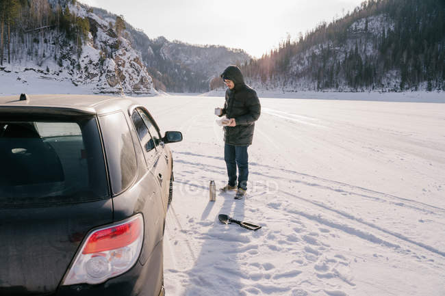 Біля автомобіля на сніговій дорозі поміж гір Сибіру (Росія) видніється чоловічий мандрівник з чашкою та термосом. — стокове фото